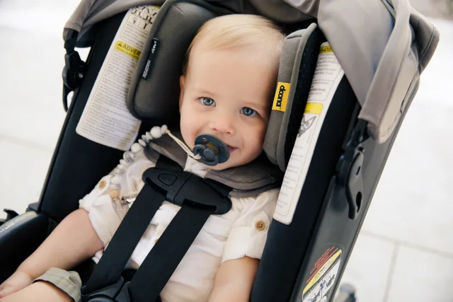 Blue-eyed blonde baby with pacifier sitting securely harnessed in a gray and black Doona infant car seat stroller