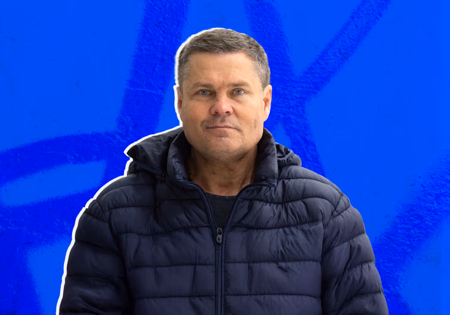 Man with short gray hair wearing a dark blue quilted jacket standing against a bright blue background.