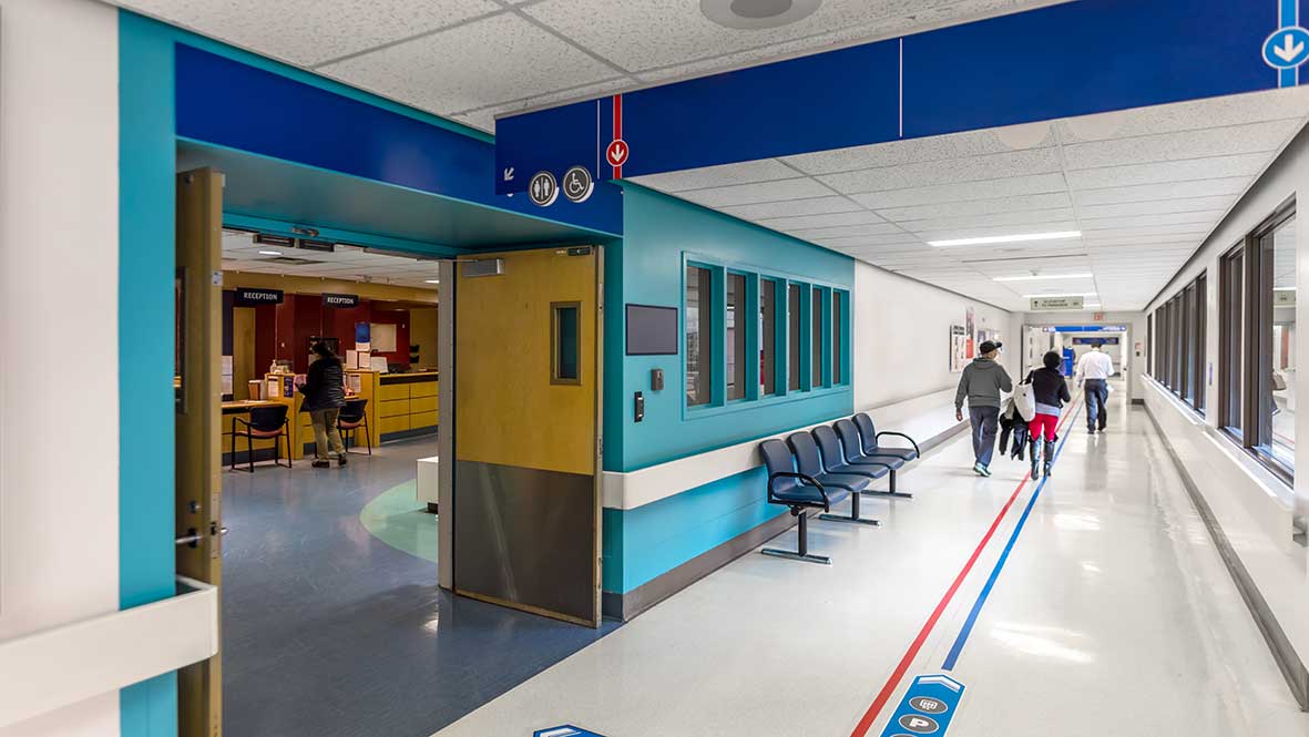 Hospital corridor with chairs along wall, people walking, and open doors to reception area on the left.
