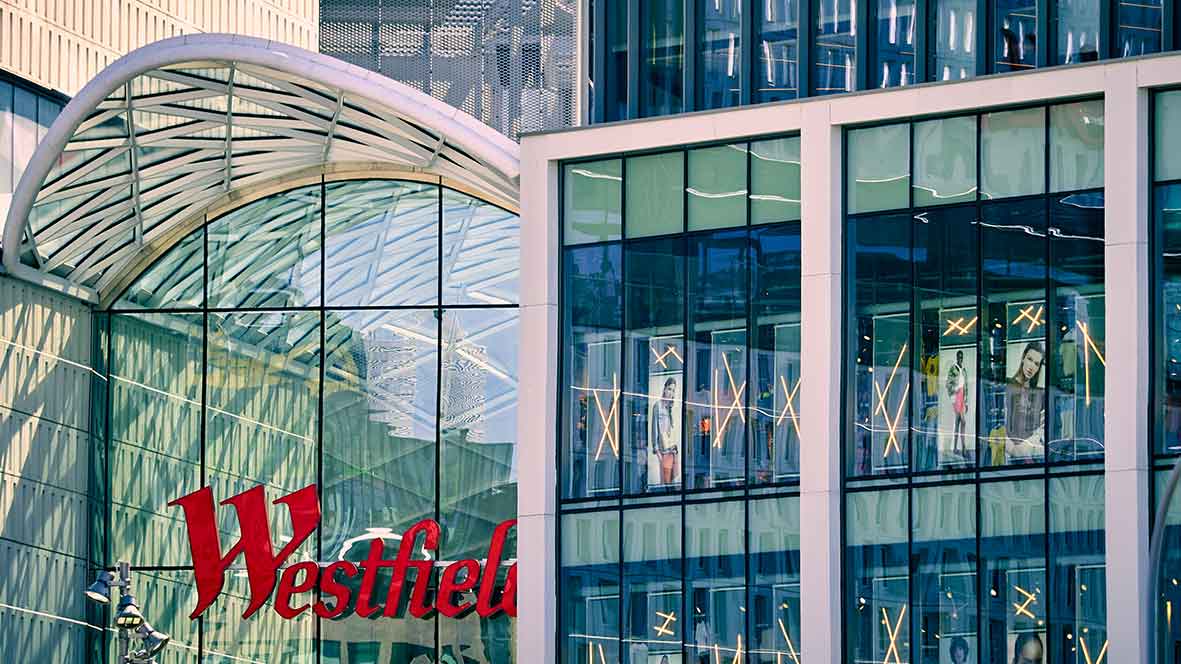 Glass facade of a Westfield shopping mall with reflections and fashion advertisements inside.