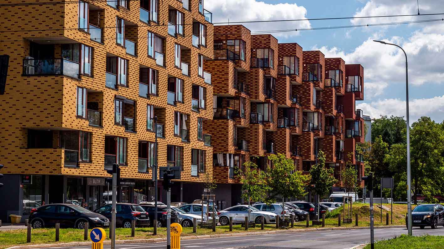 Modern residential buildings with yellow and brown brick facades and parked cars in front on a sunny day.