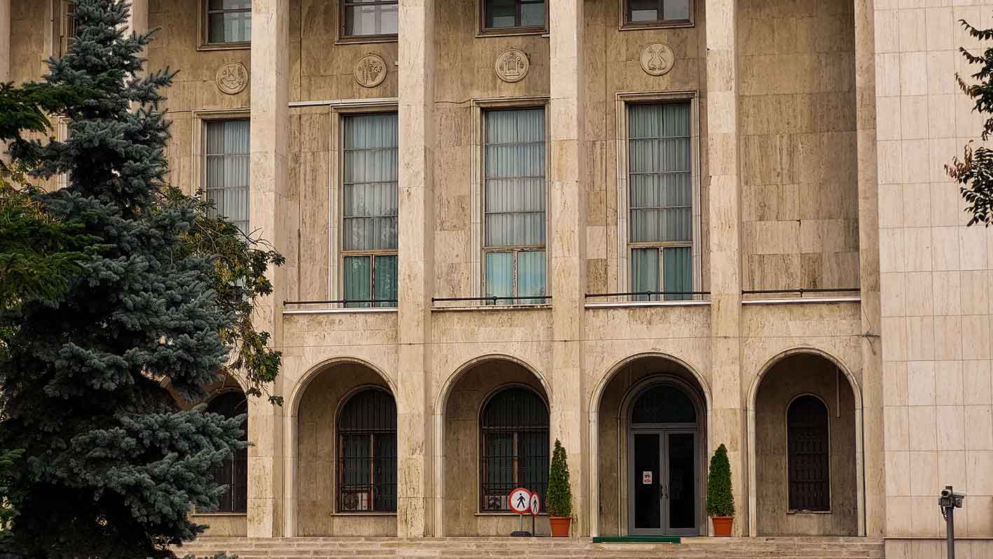 Old stone building facade with columns, tall windows, arched doorways, two potted plants, and trees nearby.