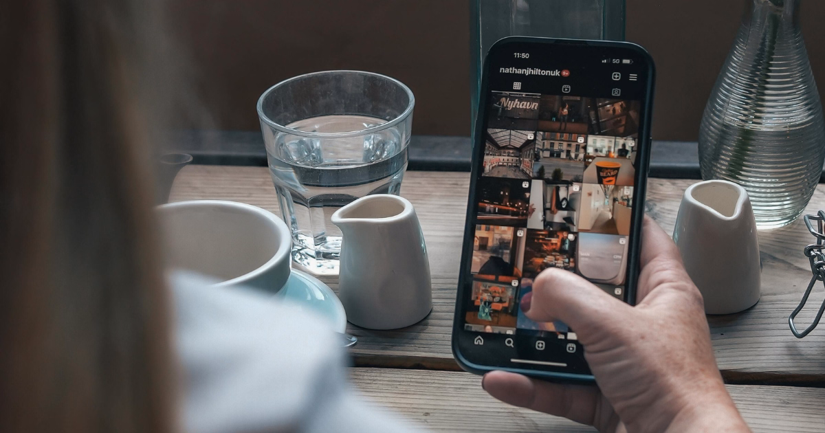 Social media manager scrolling through Instagram content on a smartphone while working at a café table with a coffee cup and glass of water, reviewing social media posts and engagement.