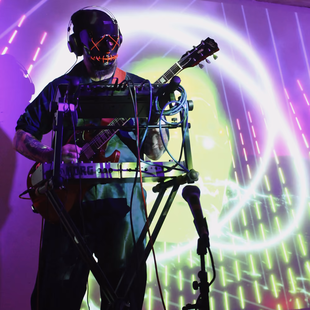 Musician wearing a black mask with red LED stitched mouth and eyes, playing electric guitar behind electronic equipment and microphone stand, with green and purple neon abstract light projections in the background.