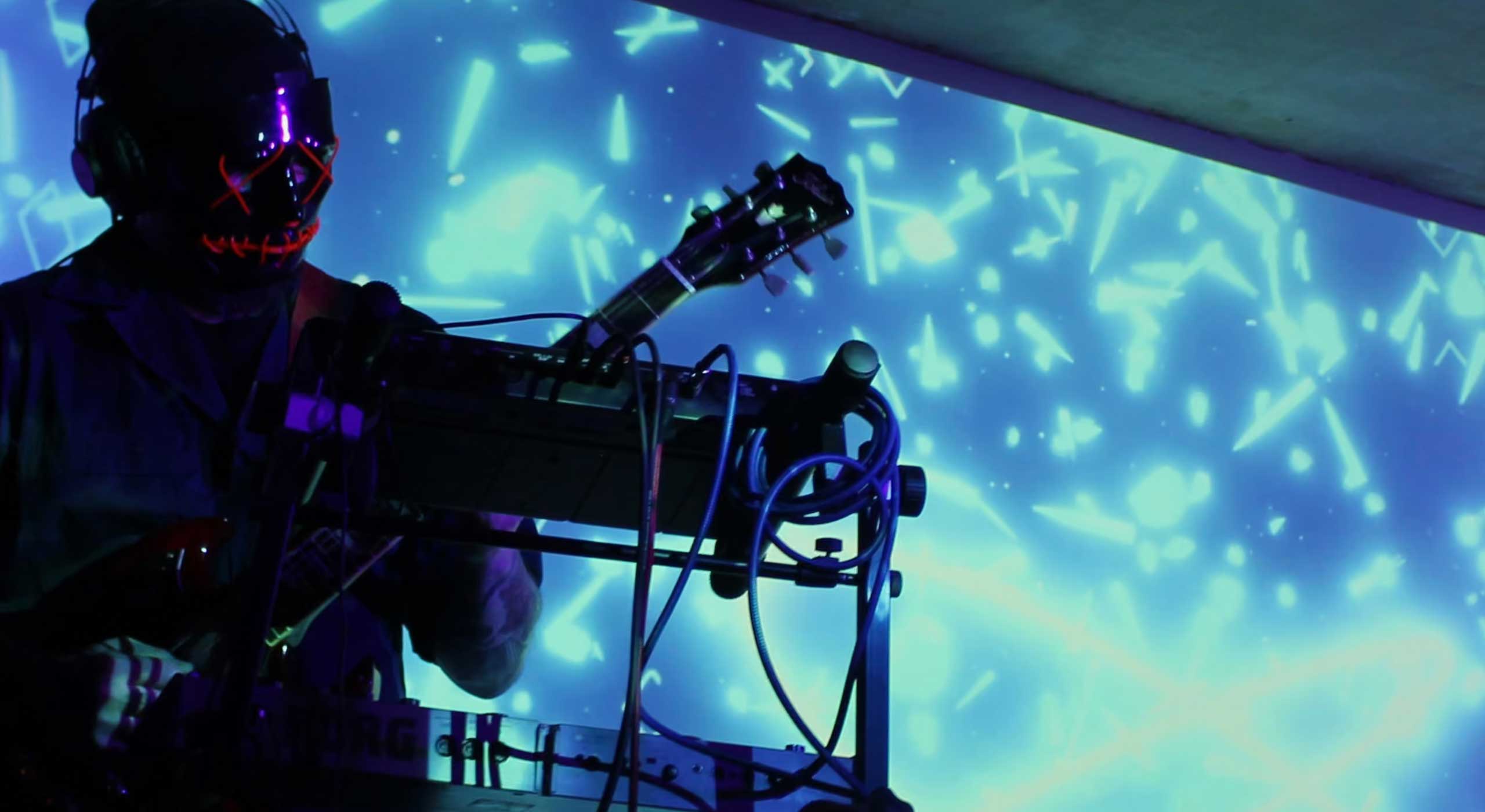 Musician wearing LED-lit mask performing with guitar against a blue abstract light background.