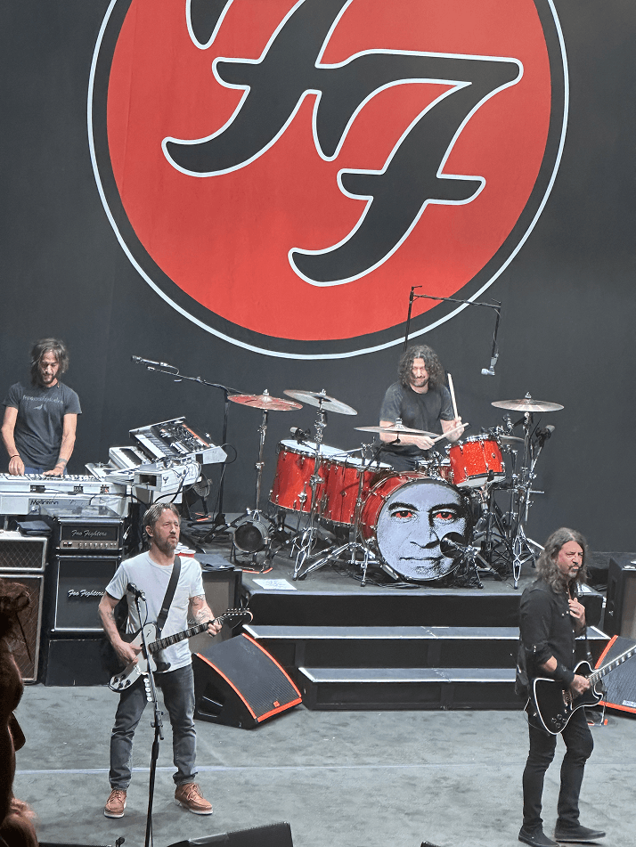 Rock band Foo Fighters performing live on stage with lead singer on guitar, drummer behind red drum set featuring a face illustration, keyboard player, and a large Foo Fighters logo in the background.