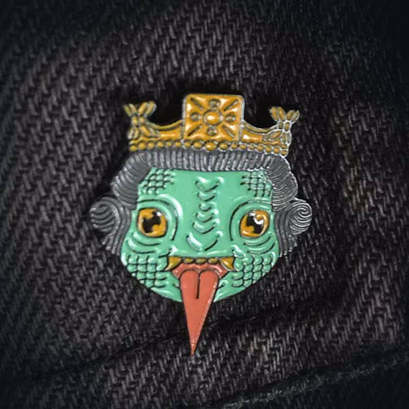 Enamel pin featuring a stylized green reptilian face with orange eyes, gray hair, a golden crown, and a long red tongue sticking out.