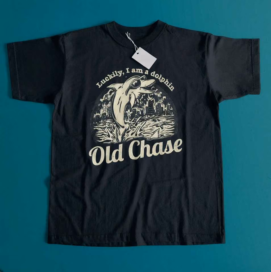Black short-sleeve T-shirt with a vintage-style graphic of a dolphin wearing sunglasses jumping out of the water in front of a city skyline and sailboat, with the text 'Luckily, I am a dolphin' above and 'Old Chase' below the illustration.