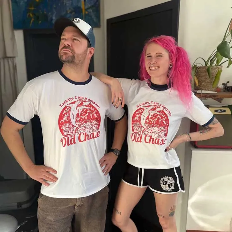 Man and woman standing side by side indoors wearing matching white ringer t-shirts with red graphic of a dolphin jumping out of water and the text 'Luckily, I am a dolphin Old Chase'. The man wears a cap and brown pants, the woman has pink hair, tattoos, and black shorts.