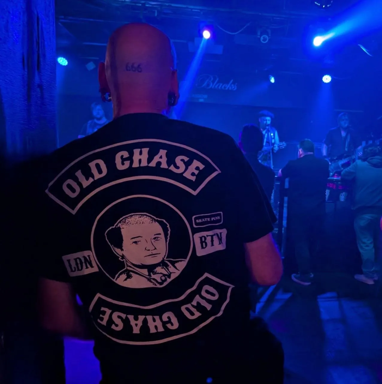 Bald man with a tattoo of the number 666 on the back of his head, wearing a black t-shirt with a graphic of a man's face and the text 'OLD CHASE' at a dimly lit concert with blue stage lighting, a band performing, and a small crowd facing the stage.