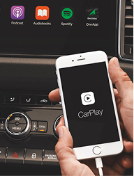 A person holding a white smartphone displaying the CarPlay logo in front of a car dashboard with control knobs and app icons for Podcast, Audiobooks, Spotify, and OneApp.