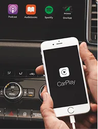 A person holding a white smartphone displaying the CarPlay logo in front of a car dashboard with control knobs and app icons for Podcast, Audiobooks, Spotify, and OneApp.