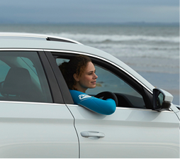 Woman with curly hair in a turquoise wetsuit sitting in the driver's seat of a white car by the beach, looking out the open window with a calm expression.