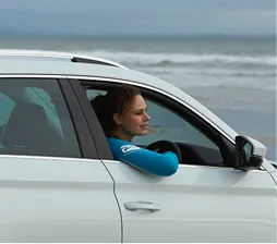Woman with curly hair in a turquoise wetsuit sitting in the driver's seat of a white car by the beach, looking out the open window with a calm expression.