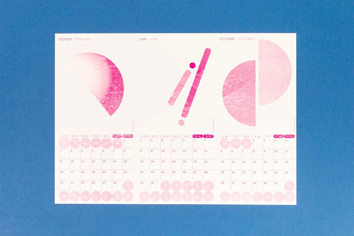Ein weißes Kalenderblatt mit pinken abstrakten Formen und den Monatsdaten für Februar, Juni und Oktober liegt auf einem blauen Hintergrund. Es wurde in Risographie gedruckt.