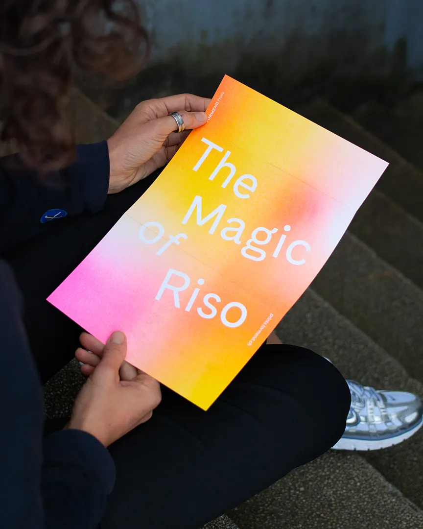Person hält ein farbigen Riso-Gutshein mit dem Text ‚The Magic of Riso‘, die Farben verlaufen von Gelb zu Pink.