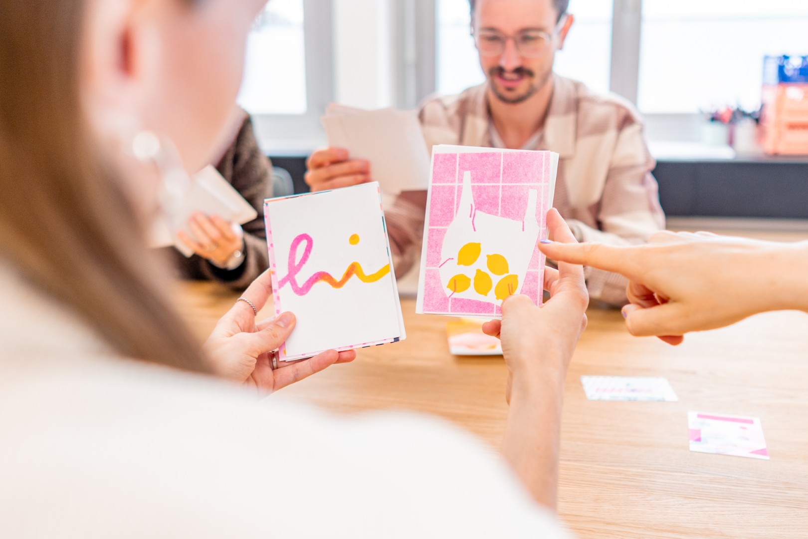 Mehrere Personen halten verschiedene Riso-Karten, eine Karte zeigt das Wort 'hi', eine andere Illustrationen von Flaschen und Zitronen.