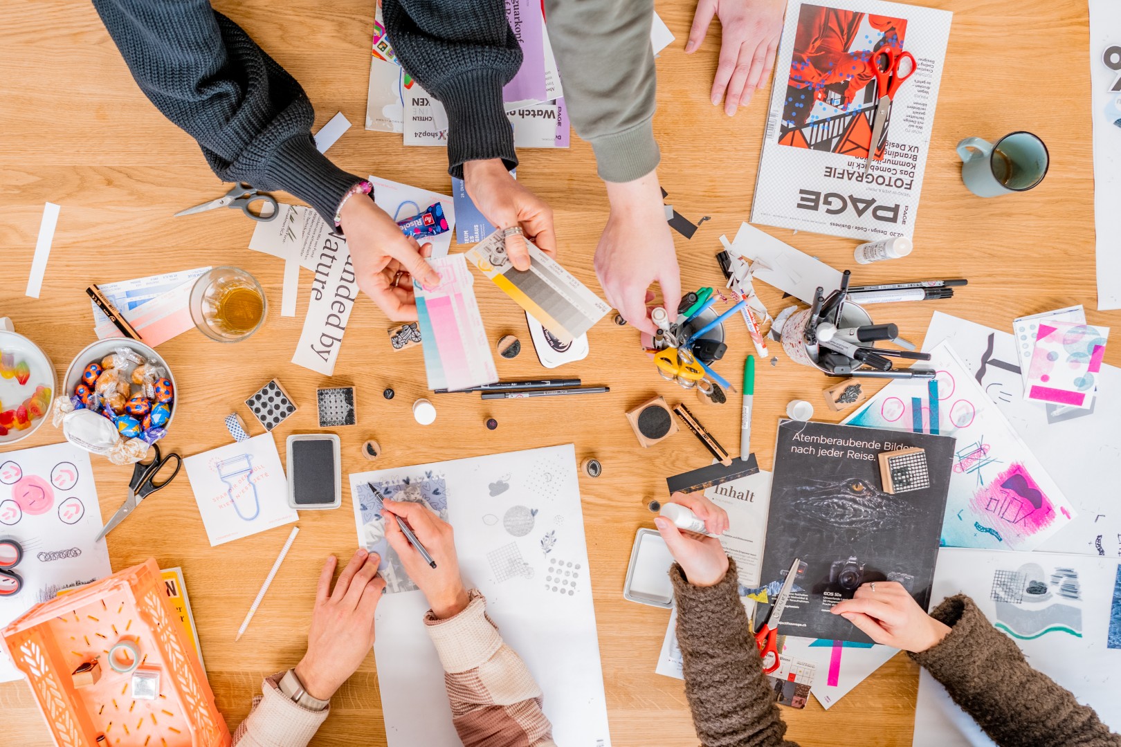Mehrere Personen arbeiten zusammen an einem Holztisch mit Bastelmaterialien, farbigen Papieren, Stiften, Scheren und Magazinen.