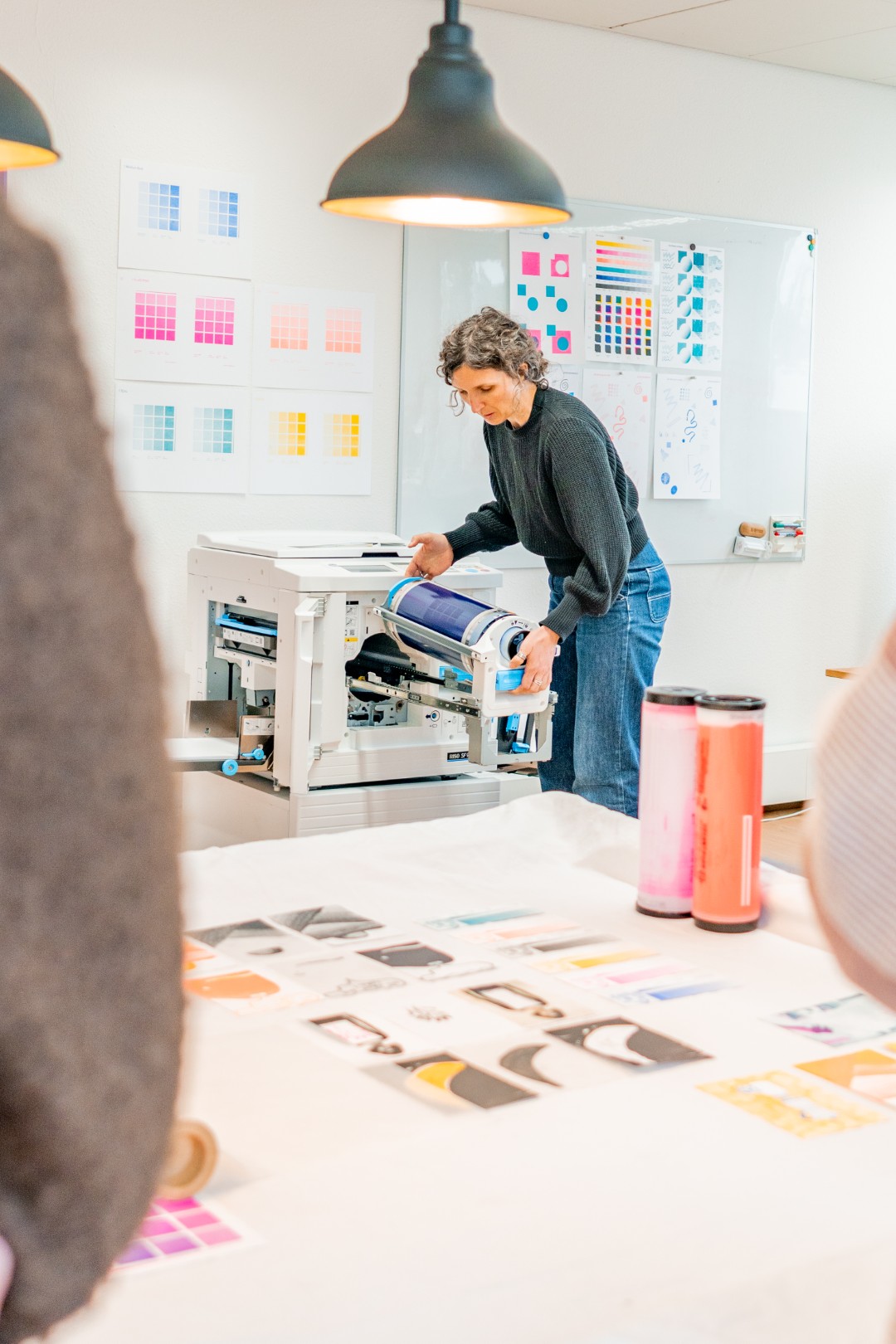 Frau arbeitet an einem Drucker und hält eine blaue Trommel, während Farbproben und Farbschemata an der Wand und auf dem Tisch im Hintergrund sichtbar sind.