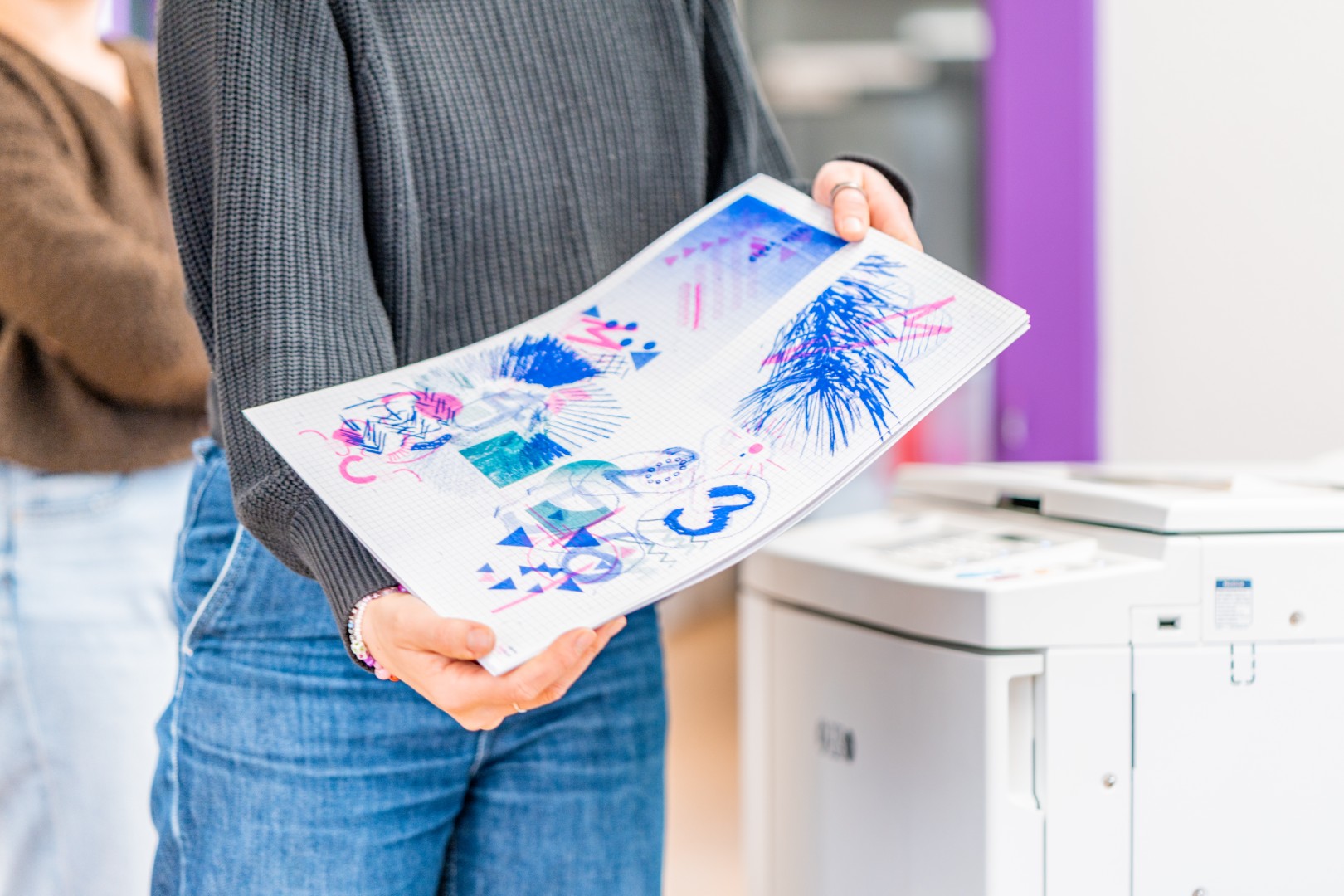 Person hält gedruckte Seiten mit farbigen abstrakten Zeichnungen neben einem Risografphen.