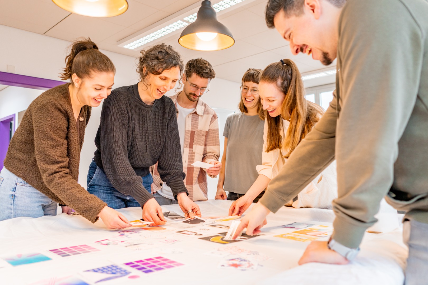 Sechs Personen stehen um einen Tisch und betrachten gespannt bunte Riso-Postkarten