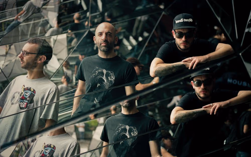 Members of the band Baja Frequencia are depicted in a creative, reflective photograph with a mirrored surface causing a kaleidoscopic effect. One member is wearing a grey T-shirt with a playful skull design, while another dons a black T-shirt with a dog print.