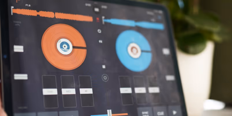 Close-up of an iPad screen displaying the Cross DJ app, with two virtual turntables in orange and blue. The app interface includes waveforms, EQ controls, and play/cue buttons, representing a digital DJ setup.