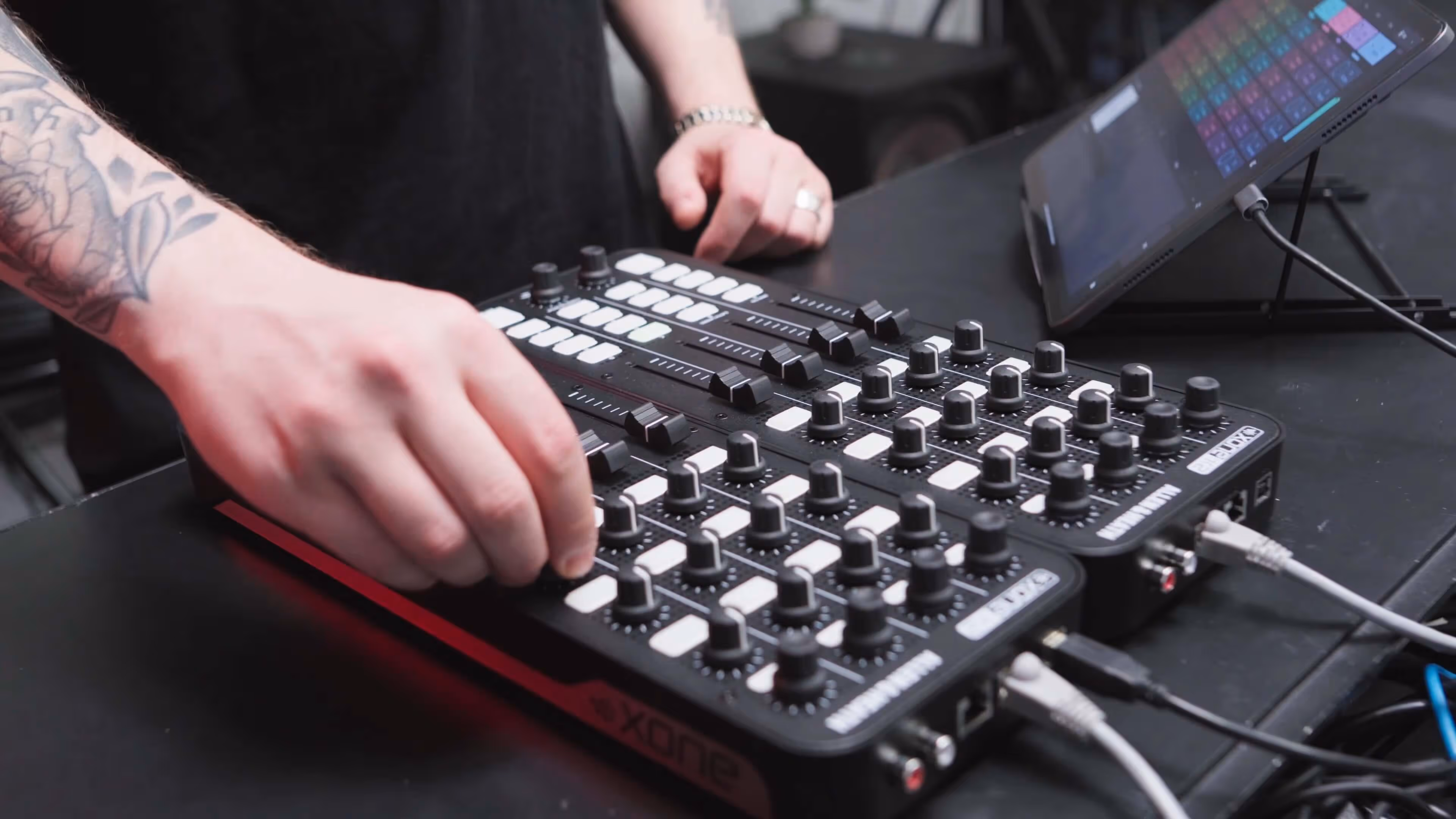 A close-up shot of a person using the Allen & Heath Xone:K2
MIDI controller, adjusting one of the knobs while a tablet running the Remixlive app is positioned on a stand nearby. The person’s tattooed arm is visible, and various cables are connected to the controller, highlighting a professional music production setup.