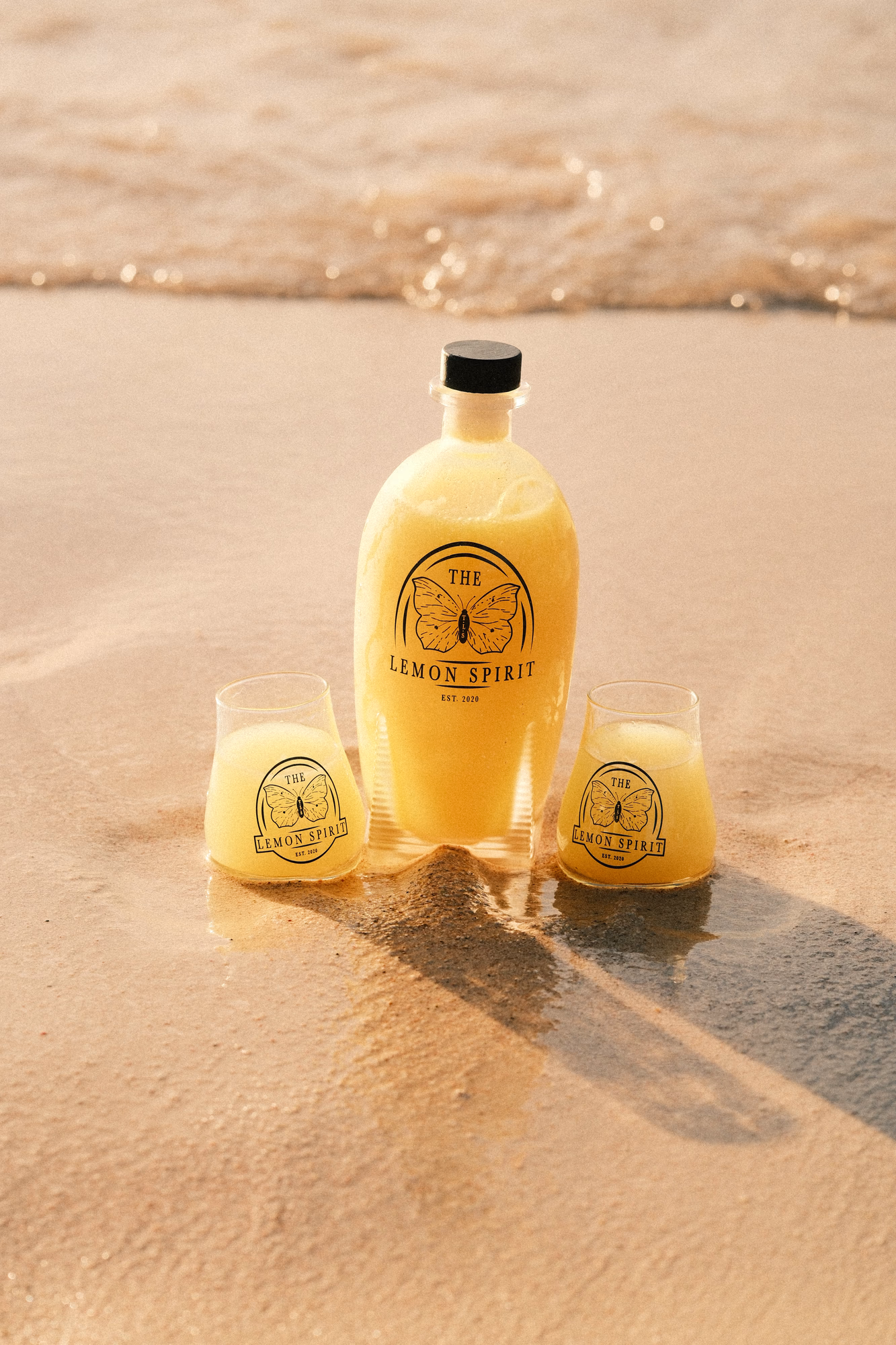 Een fles limoncello met logo van 'The Lemon Spirit' en twee met limoncello gevulde glazen in de branding op een tropisch strand.