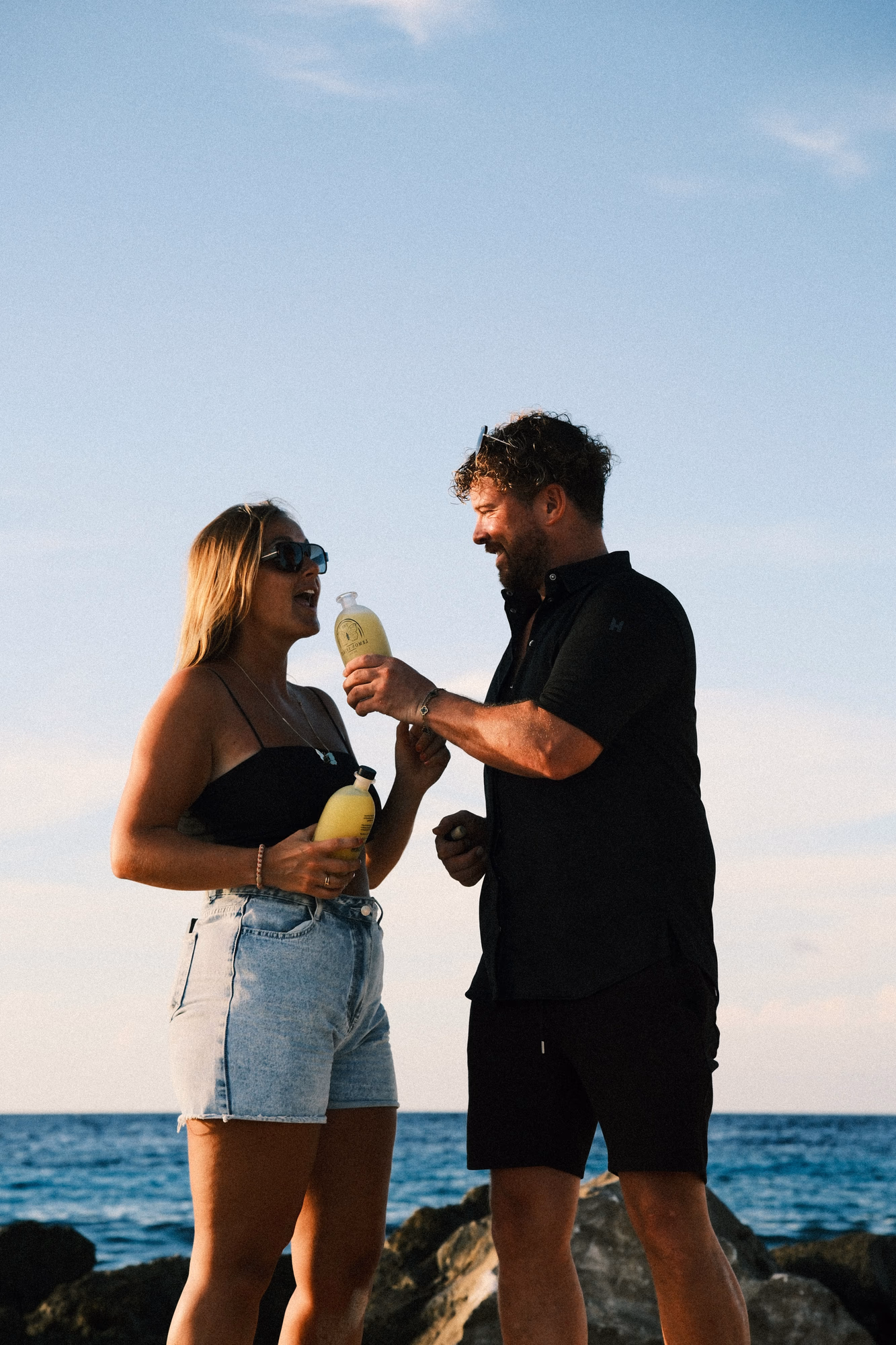 Man en vrouw staan bij de kust met blauwe lucht en rotsen terwijl ze flesjes Limoncello vasthouden en lachen.