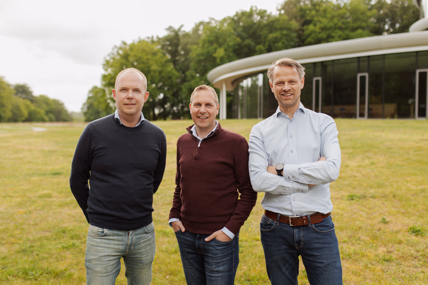 Drie mannen poseren op gras met moderne glazen gebouw en bomen op de achtergrond.