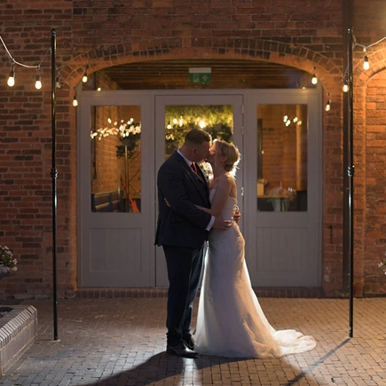 Couple embracing outside the barn doors under festoon lights at Blackbrook Barns, Lichfield, Staffordshire