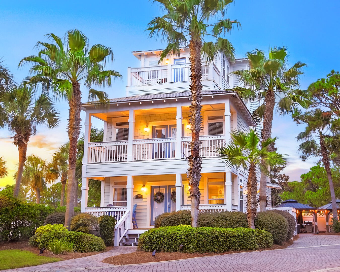 Three-story coastal vacation rental home surrounded by palm trees, featuring white siding, wraparound porches on each level, and warm lighting at sunset.