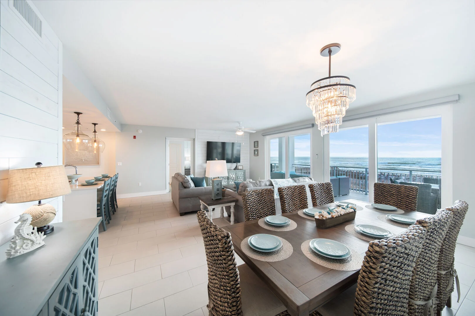Open-concept coastal condo dining and living area with wicker chairs, crystal chandelier, kitchen island, and floor-to-ceiling windows to an oceanfront balcony.