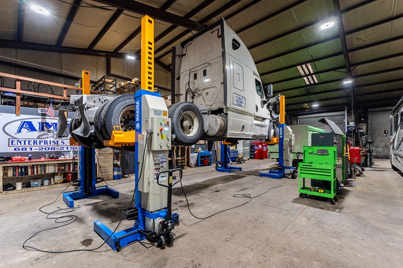 Semi-truck suspended on hydraulic lifts inside a commercial repair shop, with visible tools, equipment, and AMF Enterprises signage in background.