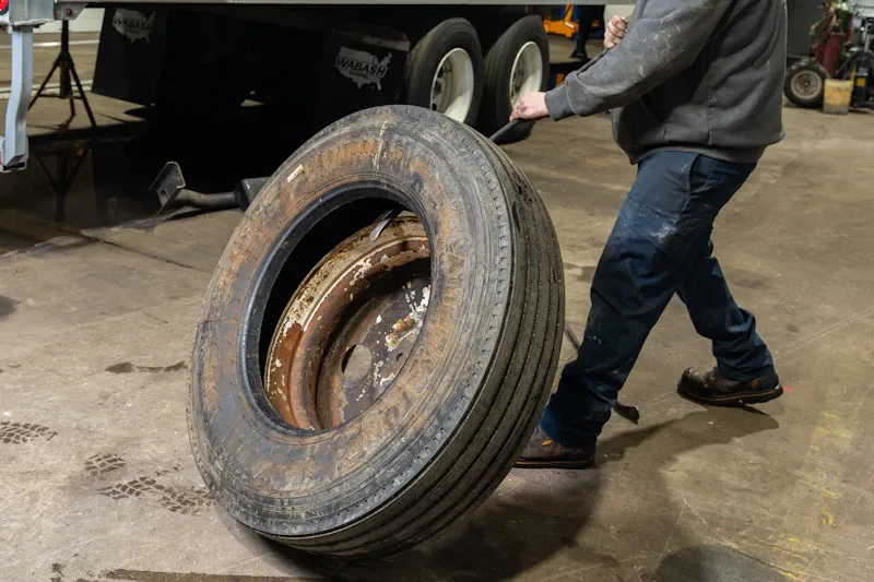 Tire service with a technician rolling a mounted semi-truck tire and wheel through a heavy-duty repair shop. Wabash mud flaps are visible.