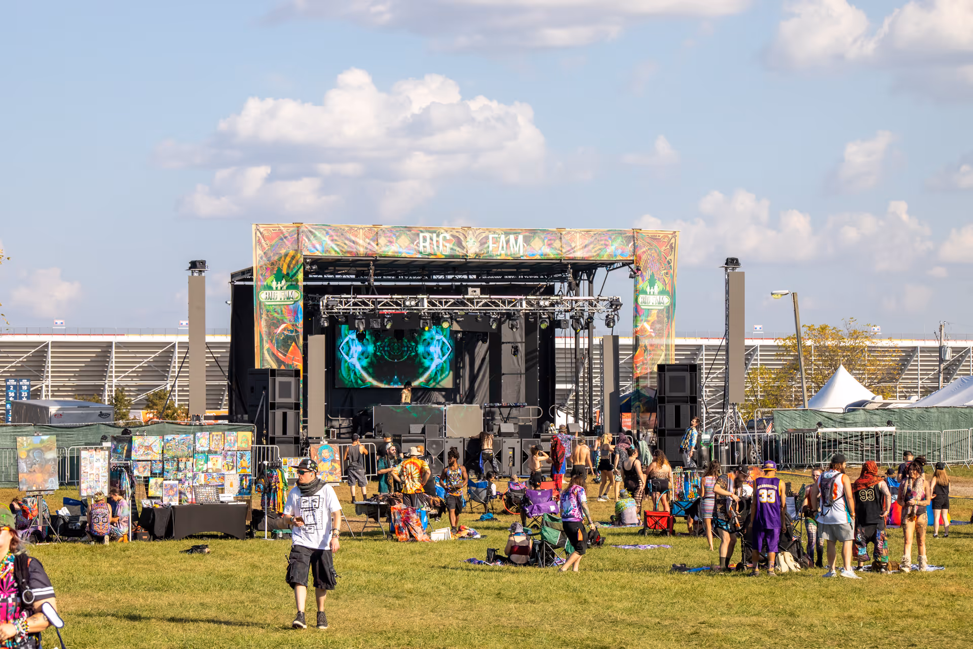 Outdoor music festival with a stage labeled Big Fam, people walking and sitting on grass, and art displayed on tables.