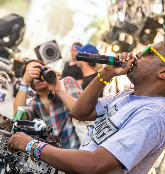 A DJ performing live at an outdoor music festival, wearing sunglasses and holding a microphone while a photographer captures the moment