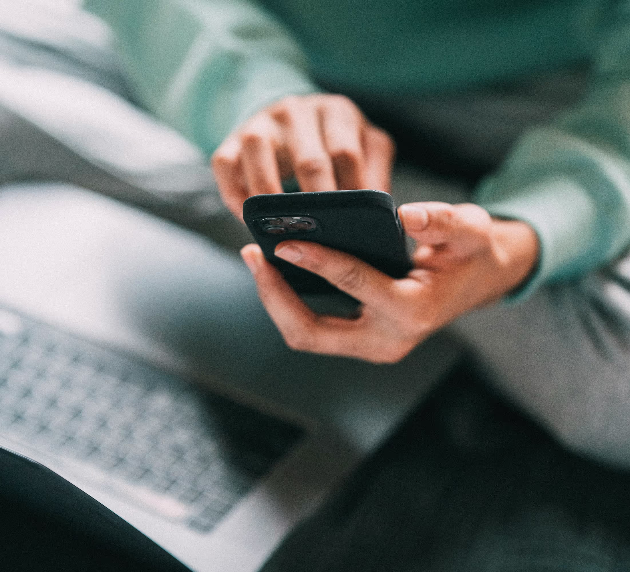 Nahaufnahme einer Person, die ein Handy benutzt, mit einem Laptop im Hintergrund.