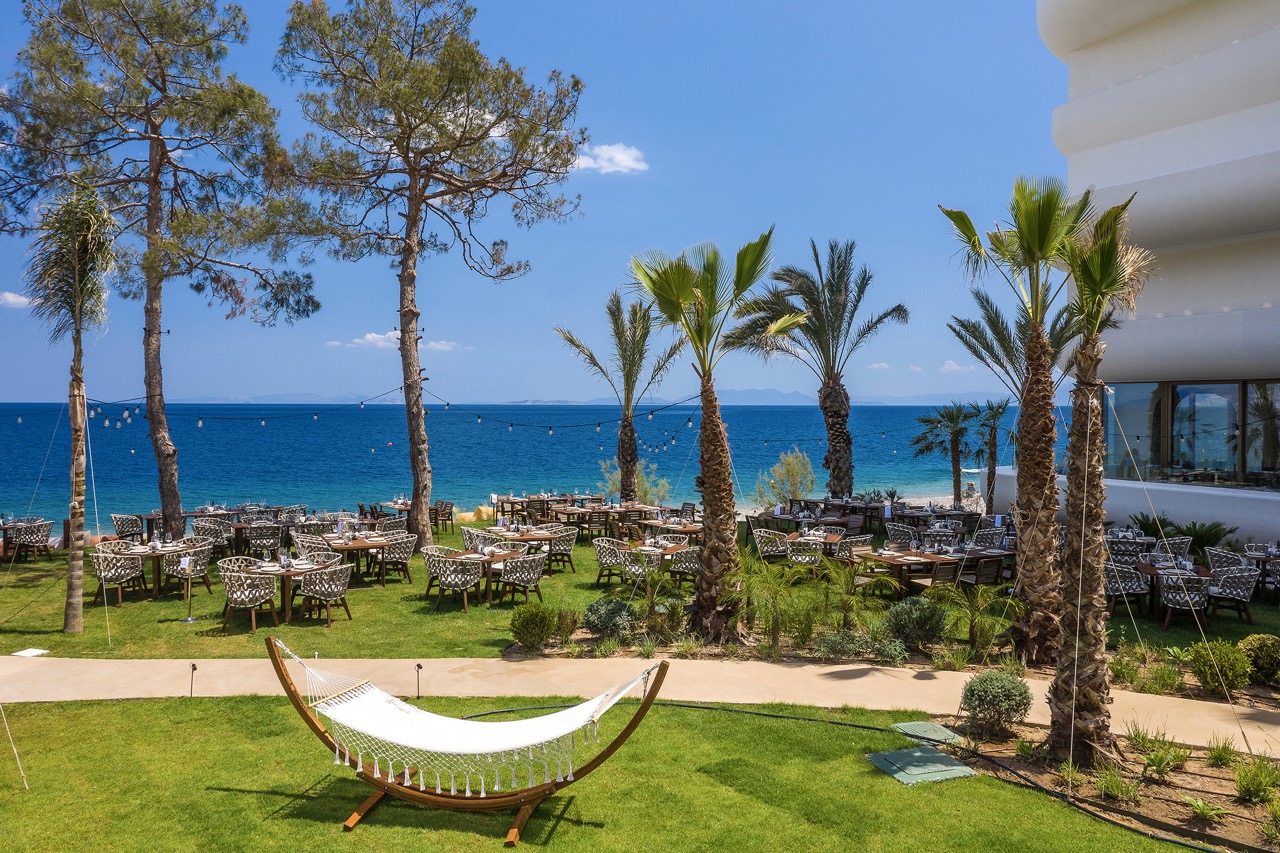 Mediterrane Taverna im Garten des Isla Brown Corinthia Resorts mit Meerblick und entspannten Sitzmöglichkeiten unter Palmen.