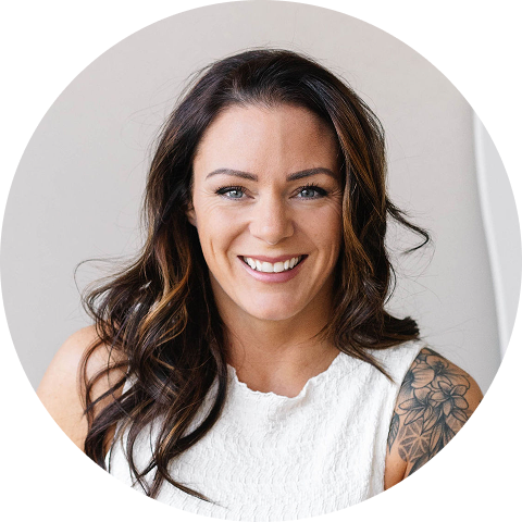 Smiling woman with long dark hair, wearing a sleeveless white top and displaying a floral tattoo on her left shoulder.