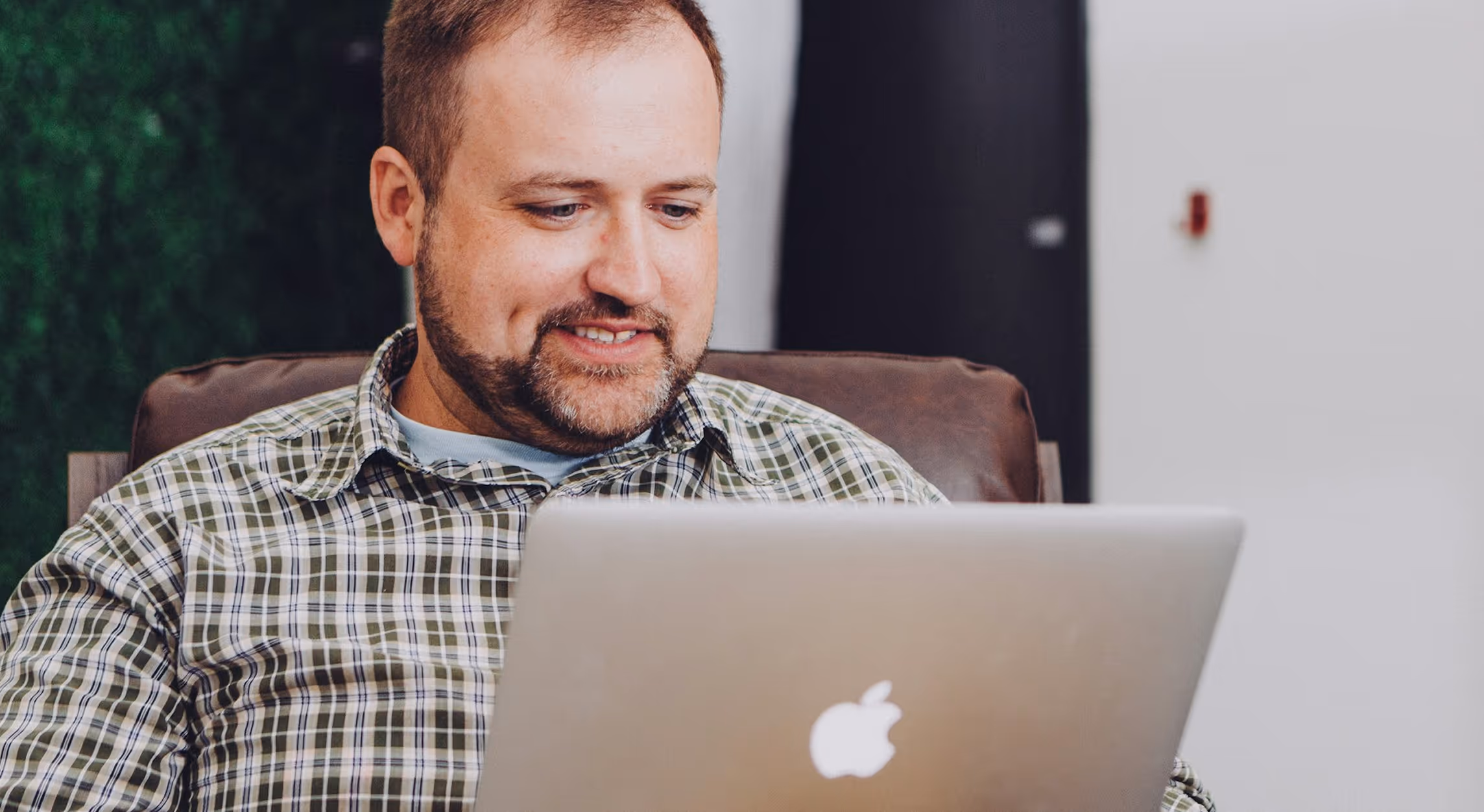 Man with beard sitting on a leather chair and smiling while looking at a silver Apple laptop.