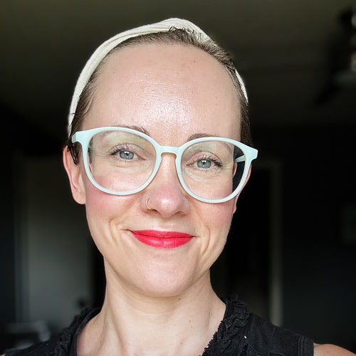 Close-up of a smiling woman wearing white-framed glasses and a white headband, with bright red lipstick.