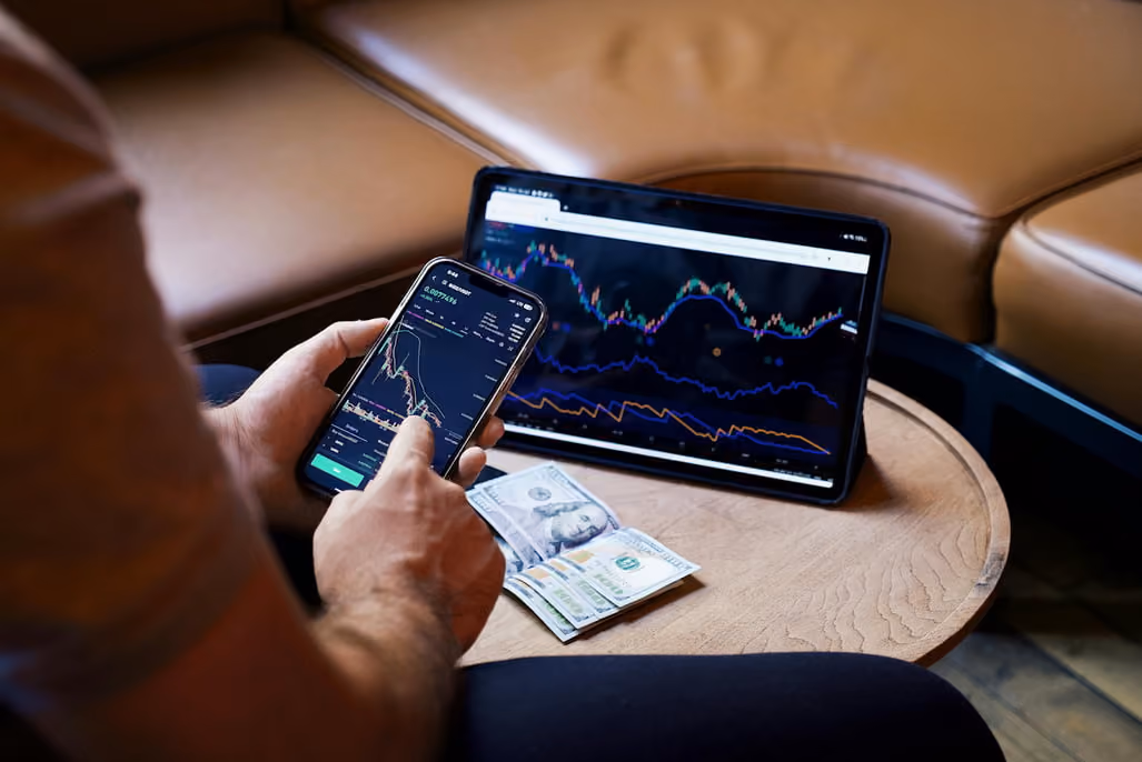 A man engaged in stock trading, using both a smartphone and a laptop for market analysis and transactions.