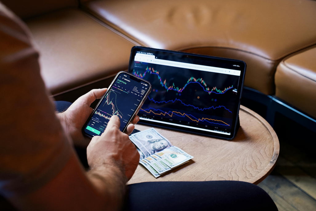 A man engaged in stock trading, using both a smartphone and a laptop for market analysis and transactions.