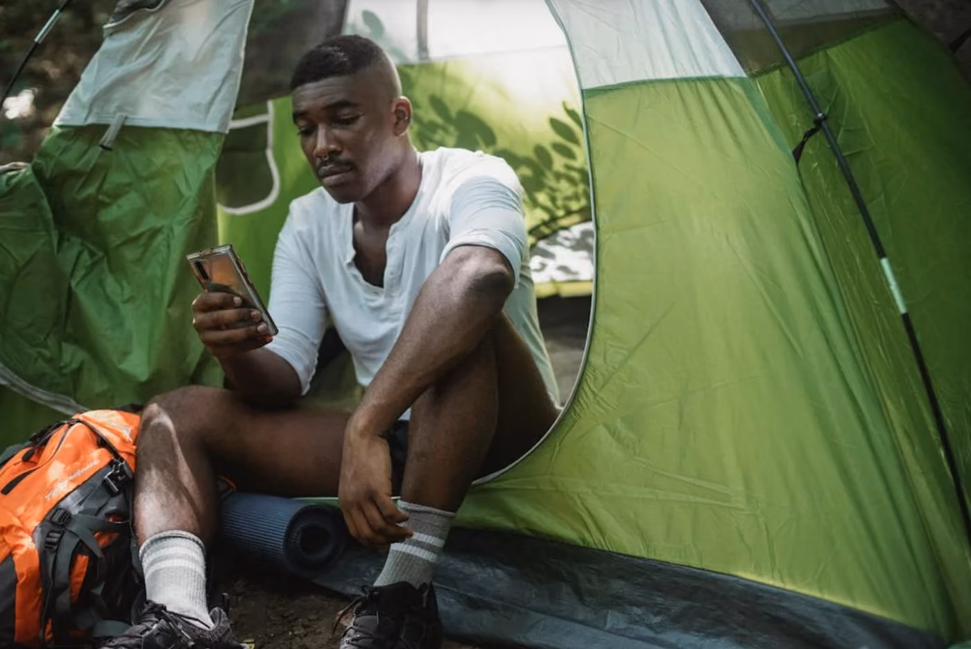 A man seated inside a tent, focused on using his smartphone, surrounded by camping gear and fabric walls.