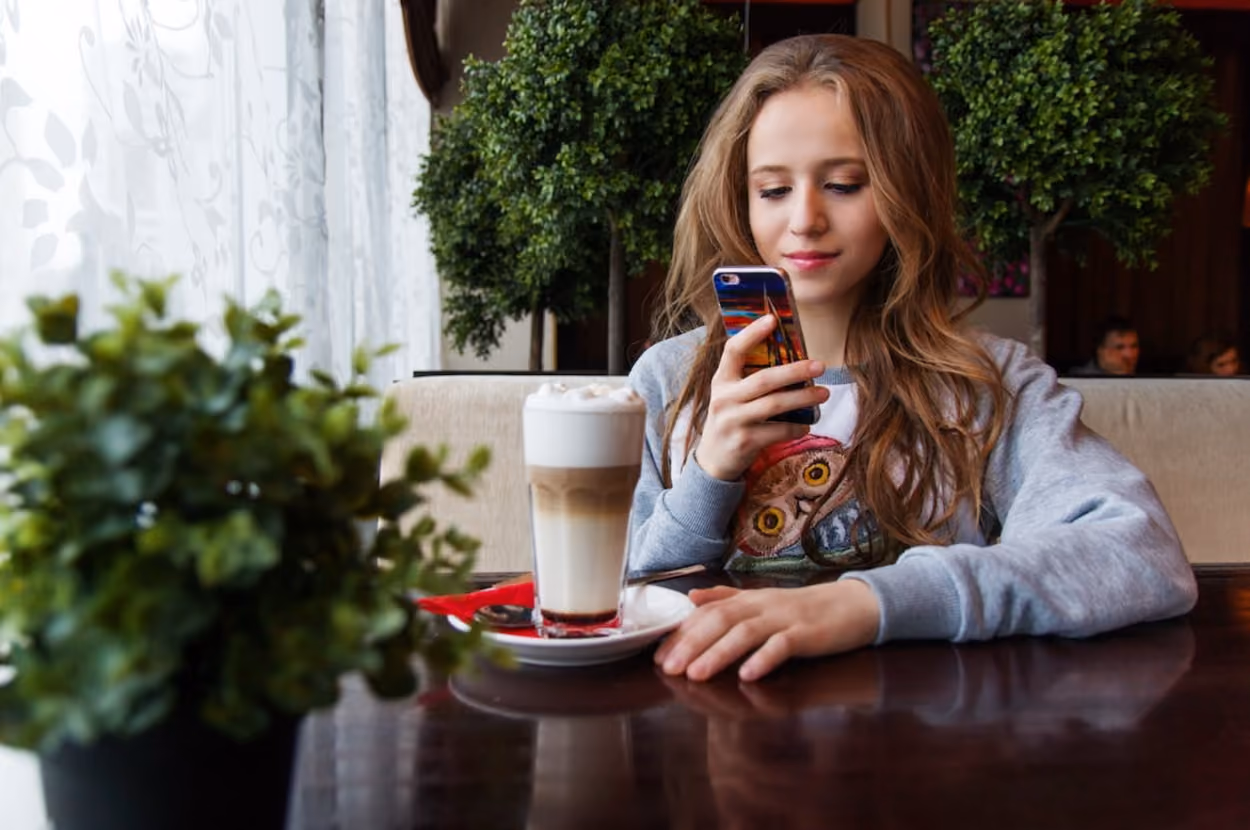 A young girl at a table, engaged with her cell phone, accompanied by a steaming cup of coffee.