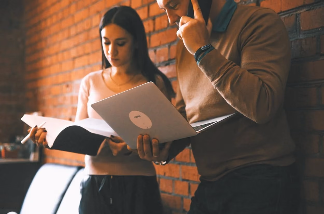 A man and woman stand beside a brick wall, each using a laptop, engaged in a focused discussion or work.