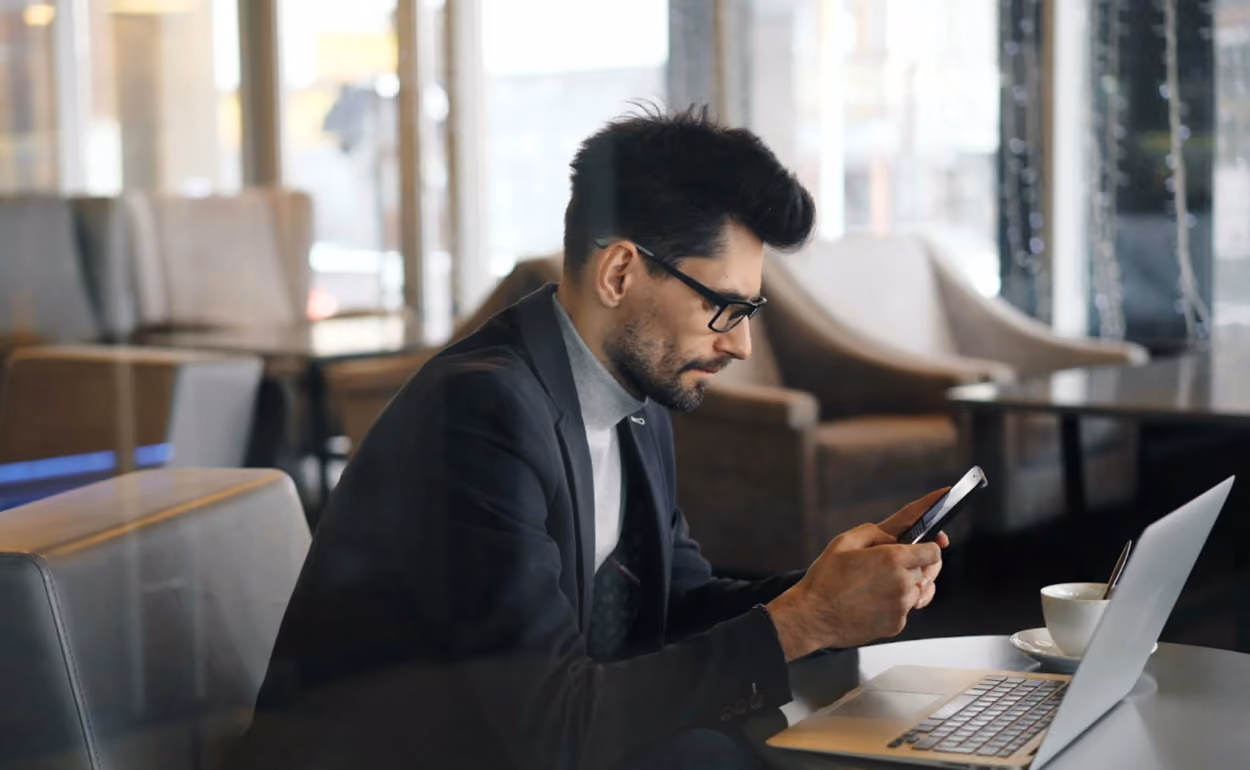 A man in a suit sits at a table, engaged in a phone conversation while focused on his device.