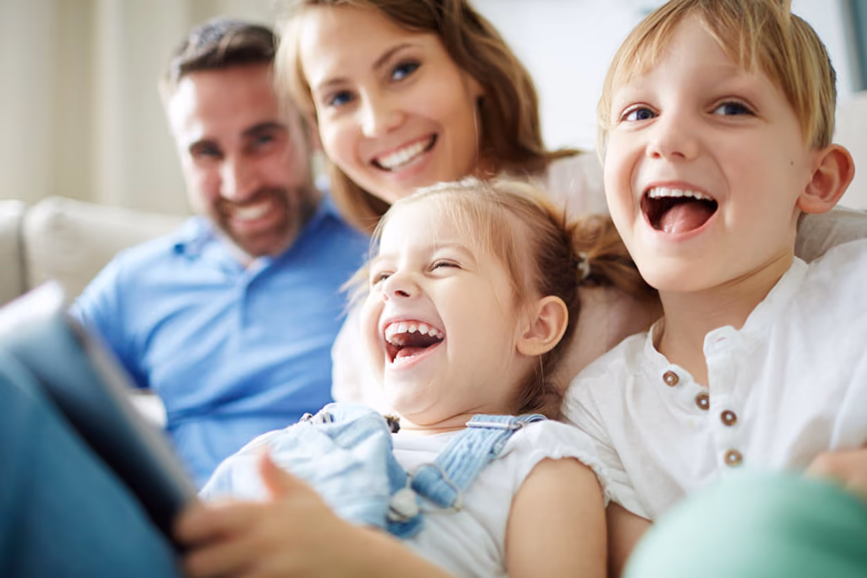 A family of four sits together on a couch, engaging with a tablet, sharing smiles and enjoying quality time.